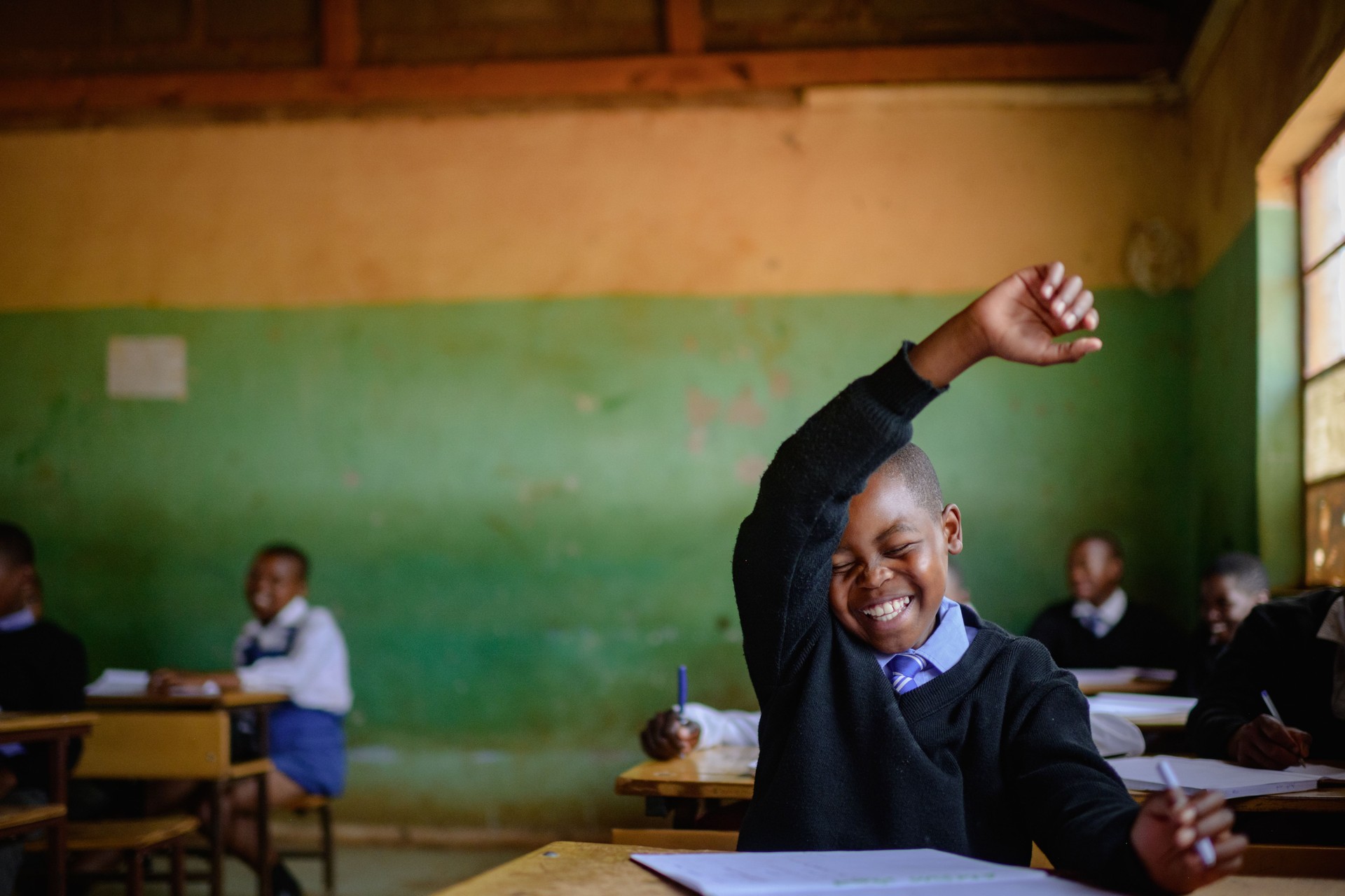 Cute Shy Schoolboy in uniform sitting in classroom hand raised smiling embarrassed